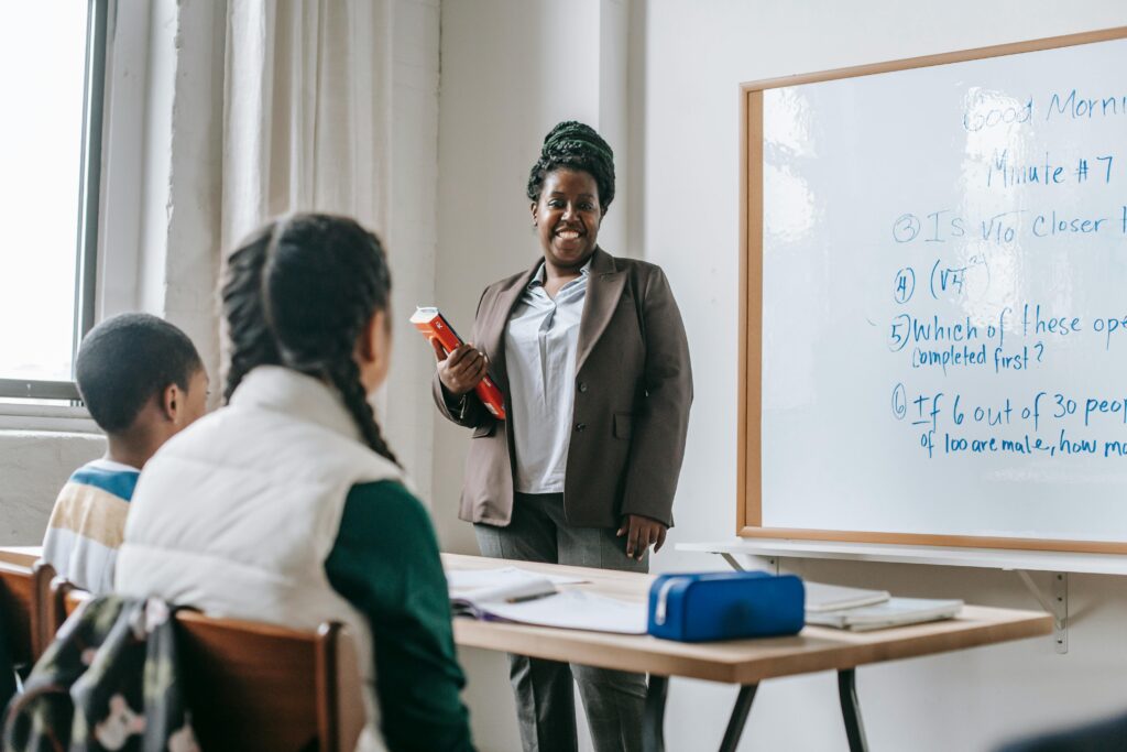Black female in teacher out standing in front of a classroom with two students in the scene smiling
